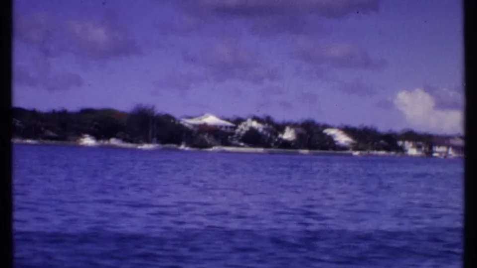 1969 Bahamas shoreline viewed from the water, with white waterfront structures, palms, and a partly cloudy sky.