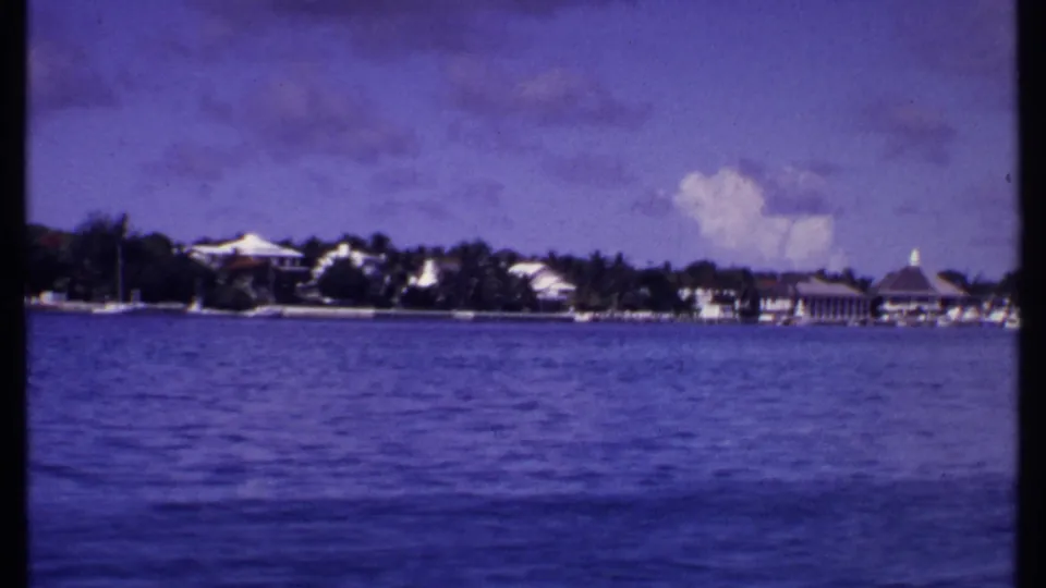 1969 Bahamas home-movie view across blue water toward a low waterfront with palms, white buildings, and small boats.
