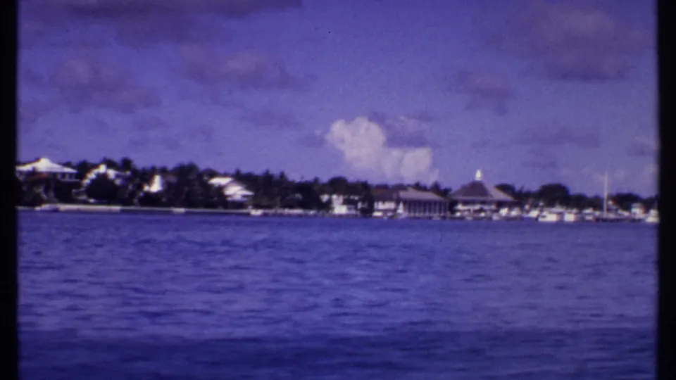 1969 Bahamas coastal view from the water, with small boats near a palm-lined shore and low white buildings beyond.