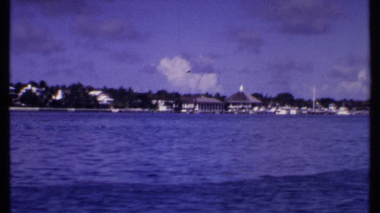 Blue ocean facing a distant waterfront with low buildings, boats, trees, and a domed roof under clouds.