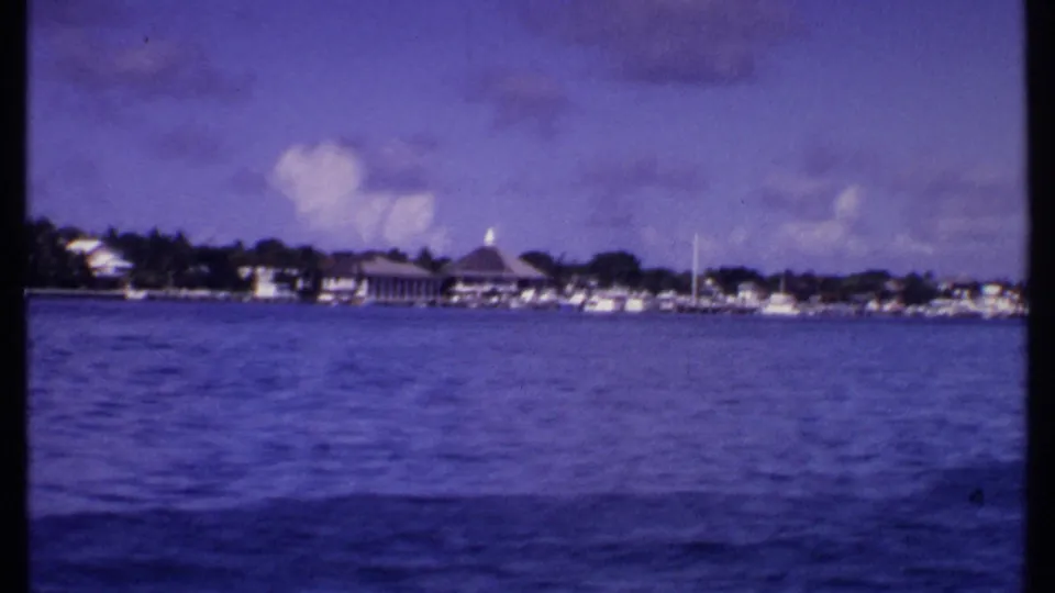 1969 Bahamas shoreline seen from offshore, with rippling water, low coastal buildings, small boats, trees, and a domed roof.