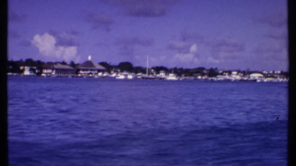 1969 Bahamas boat POV showing a coastal row of buildings, a domed roof, moored boats, trees, and blue water in the foreground.