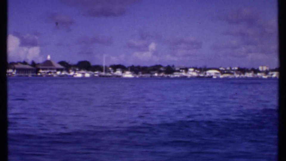 1969 Bahamas waterfront from a moving boat, with open ocean foreground, distant houses, boats, trees, and a domed structure.