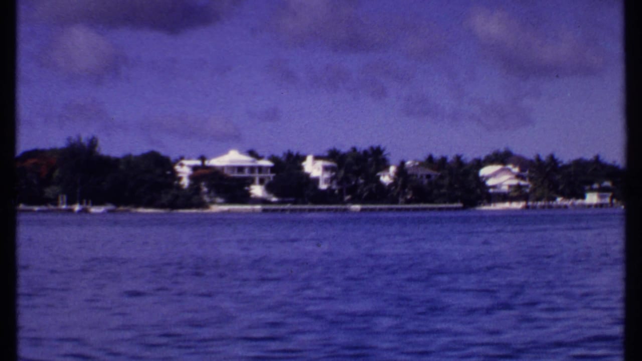 Calm water facing a distant shoreline with white houses, palms, and cloudy sky.