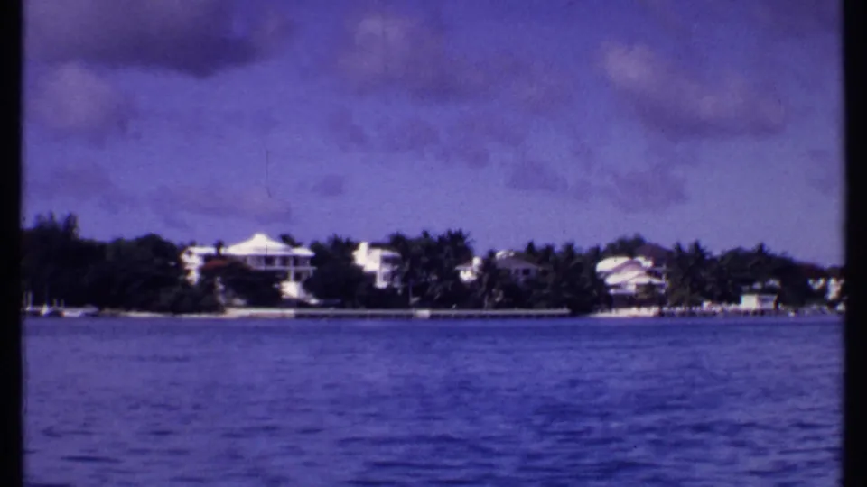 1969 Bahamas waterfront view with blue water in the foreground, white houses among palms, and low clouds overhead.