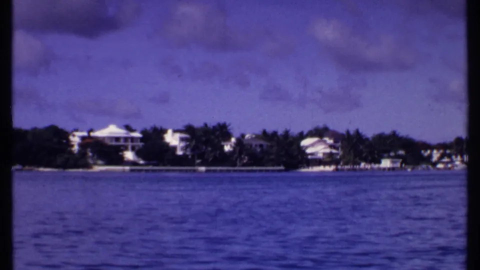 1969 Bahamas 8mm still showing a low shoreline with white homes and palms viewed from across the water.