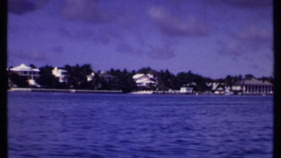 1969 Bahamas frame with calm water, distant white shoreline buildings, palms, and a columned structure at the far right.