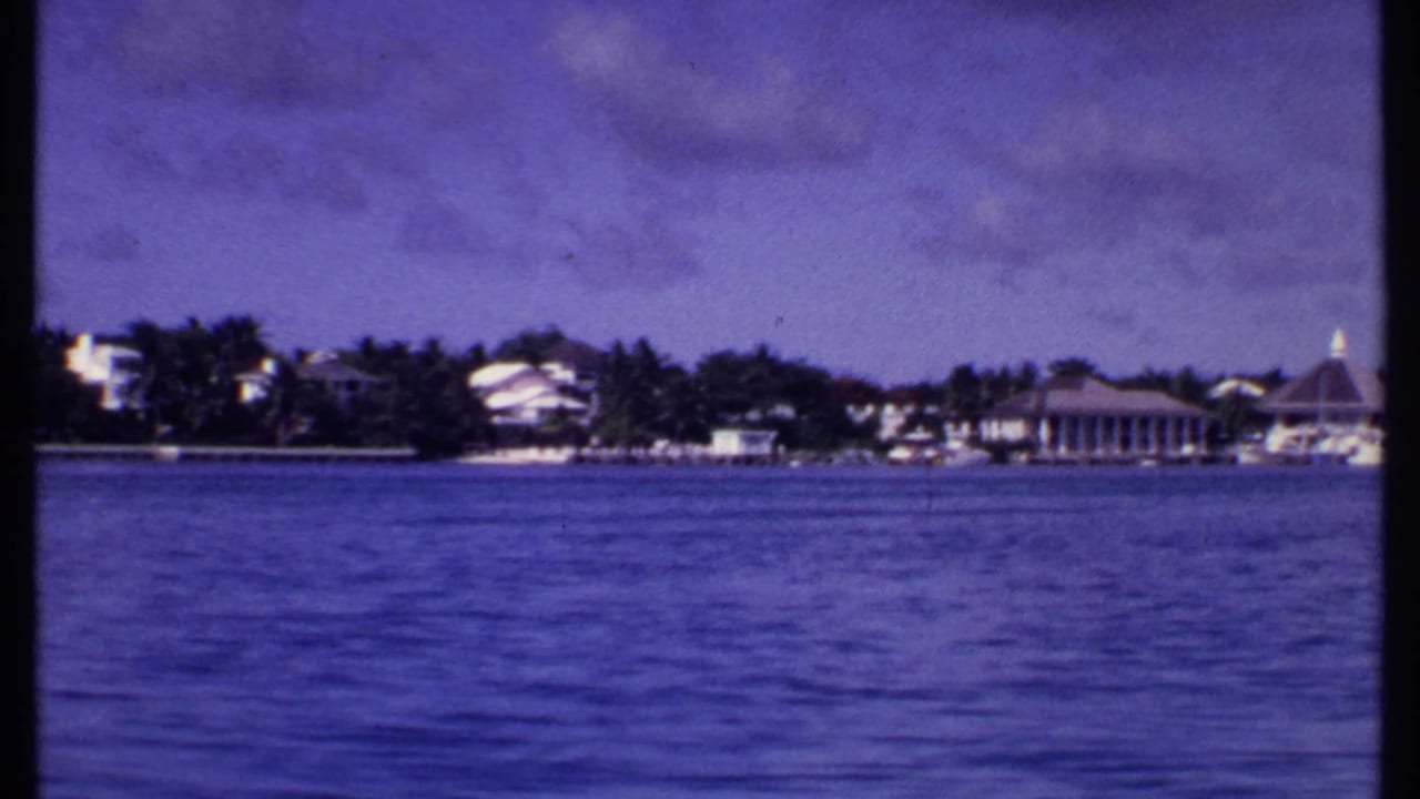 1969 Bahamas 8mm frame showing blue water, distant shoreline buildings, trees, and a pointed-roof pavilion from a moving boat.