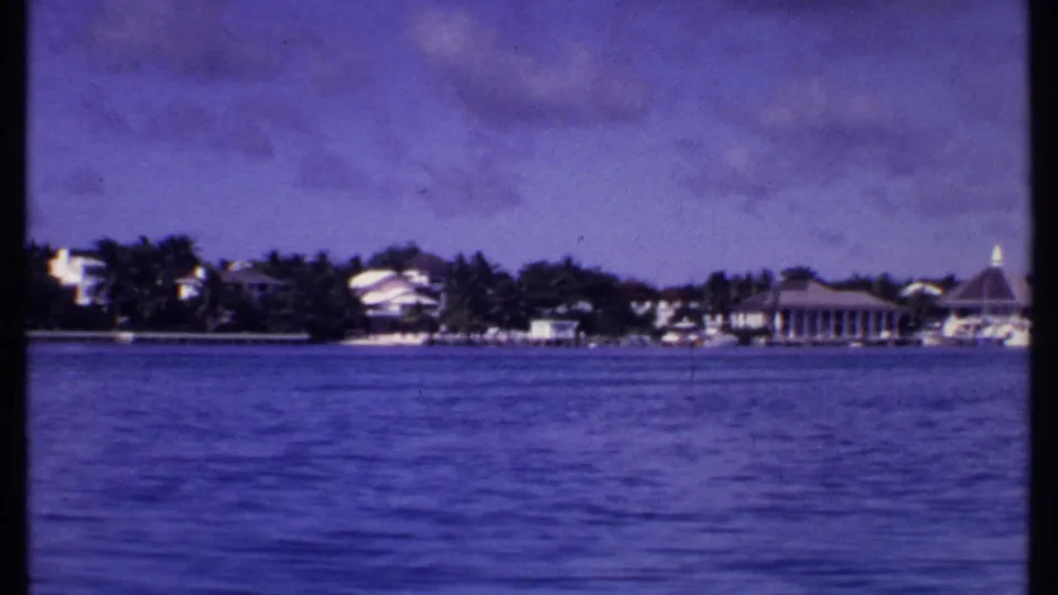1969 Bahamas 8mm frame showing blue water, distant shoreline buildings, trees, and a pointed-roof pavilion from a moving boat.