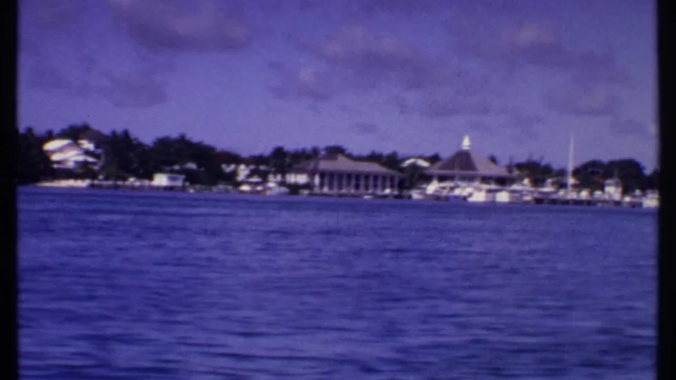 1969 Bahamas shoreline view from a boat, with docked boats, low waterfront buildings, trees, and a pavilion roof near the marina.