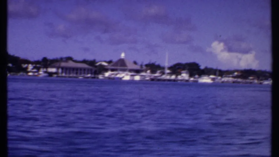 1969 Bahamas home-movie frame with calm water in the foreground, a marina, docked boats, and a pointed-roof waterfront pavilion.