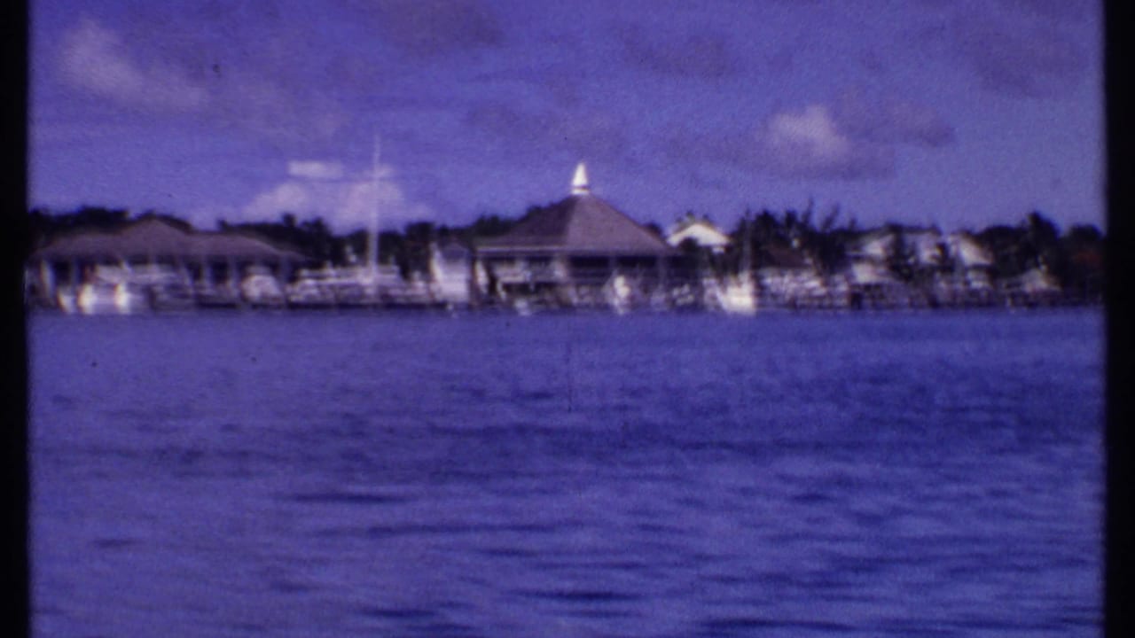 1969 Bahamas 8mm frame showing blue water, shoreline buildings, docks, palms, and a conical-roof structure in the distance.