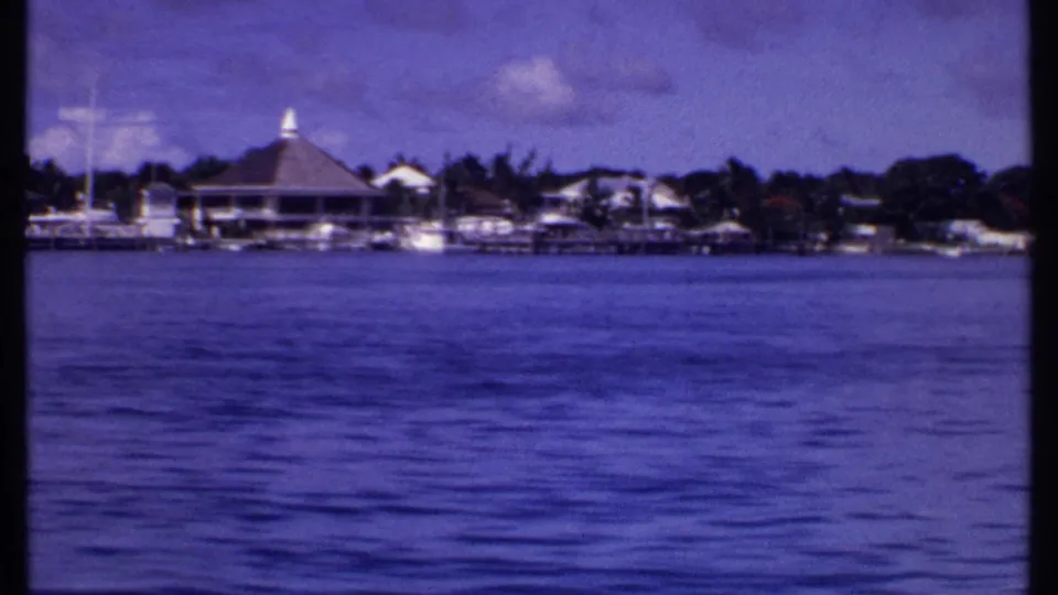 1969 Bahamas shoreline view from a moving boat, with a conical-roof building, docks, small boats, palms, and rippled water.