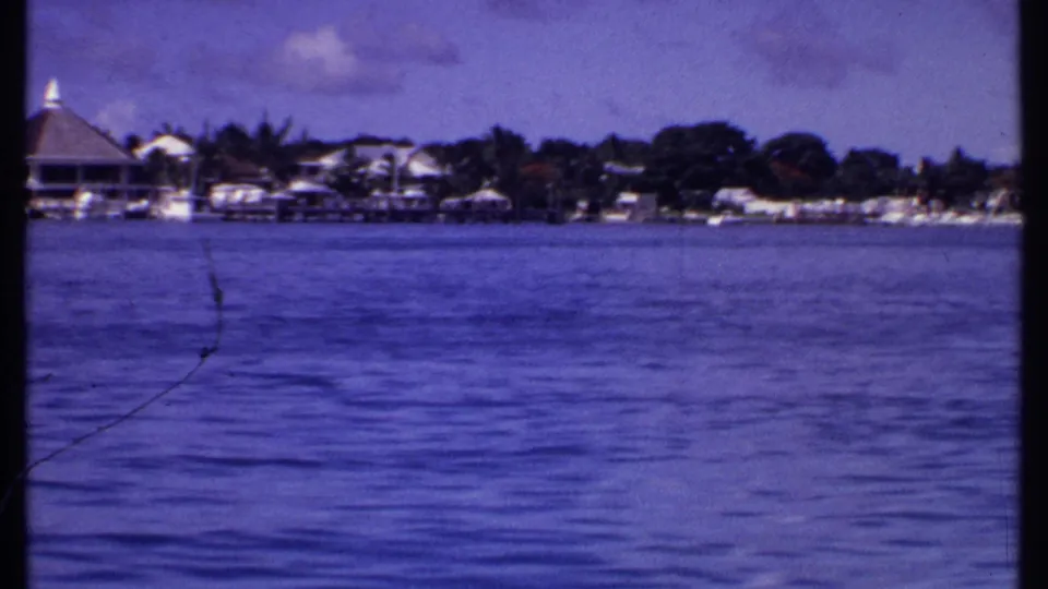 1969 Bahamas 8mm frame showing a passing view of shoreline homes, docks, palms, boats, and broad blue water.