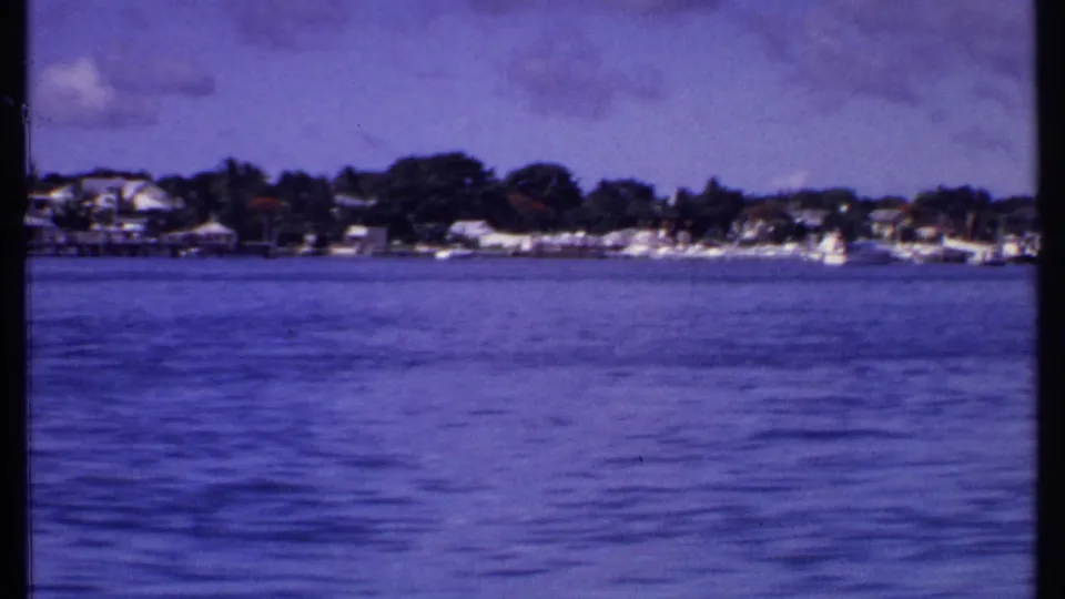 Wide 1969 Bahamas boat-view frame with low shoreline buildings, small boats, dense trees, clouds, and textured blue water.
