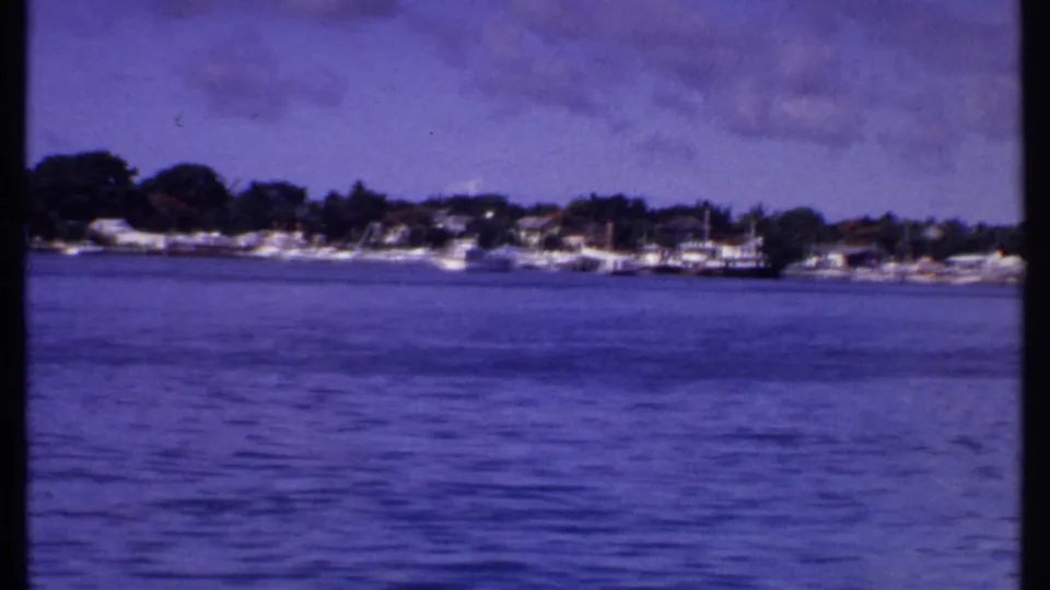 1969 Bahamas waterfront view with boats along a low shoreline, houses among trees, and a purple-blue sky above.