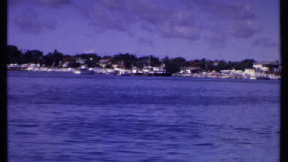 1969 Bahamas home-movie still showing rippled blue water, a distant residential coast, moored boats, and trees under clouds.