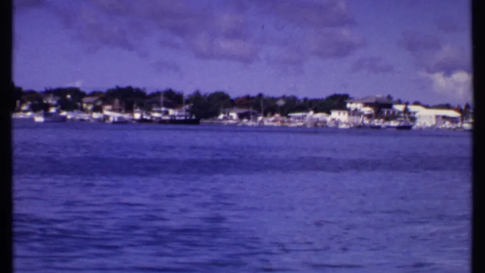 1969 Bahamas shoreline view across calm water, with boats near coastal homes and trees beneath a softly clouded sky.