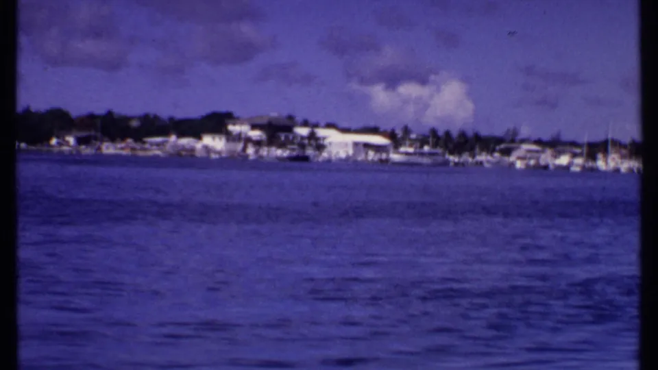 A 1969 Bahamas shoreline appears across blue harbor water, with white buildings, small boats, palms, clouds, and aged 8mm color.