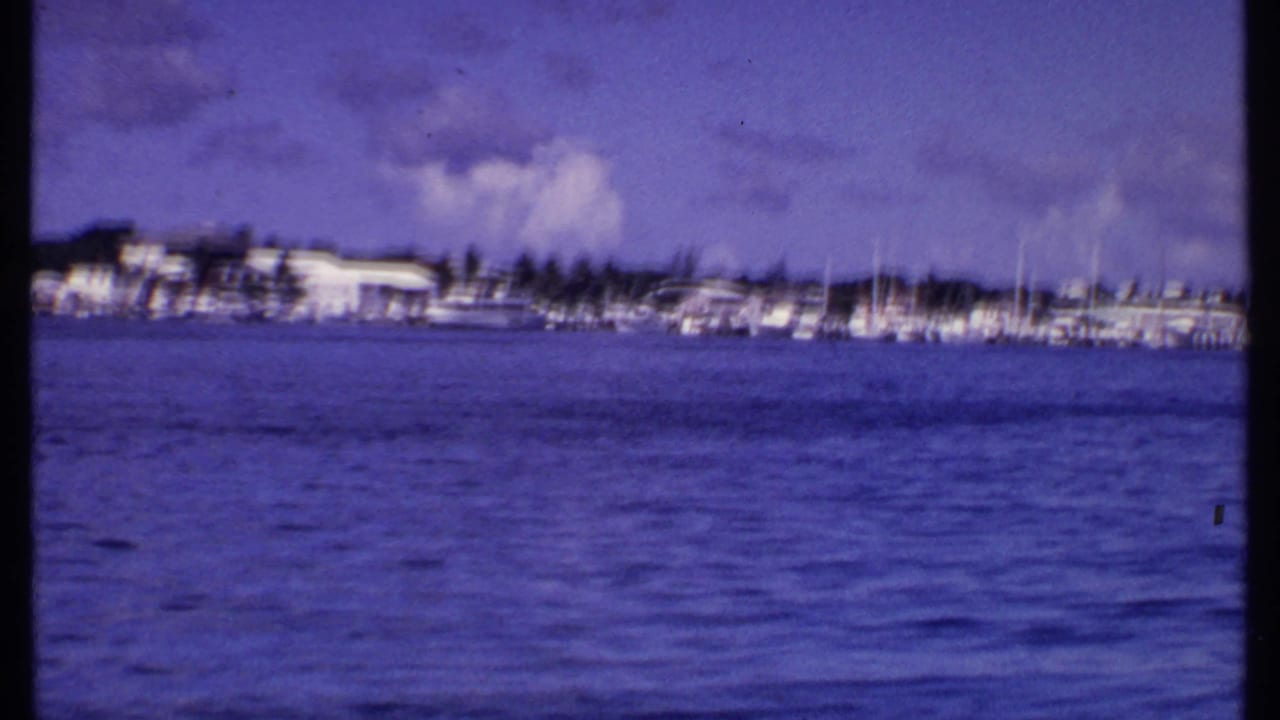 1969 Bahamas waterfront seen from the water, with docked boats, low buildings, palms, and cloudy sky in soft 8mm color.