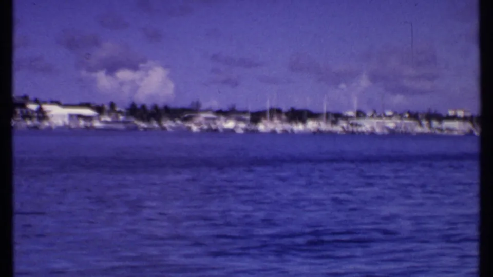 1969 Bahamas harbor view with sailboat masts, low waterfront buildings, palms, and a purple-blue 8mm sky.
