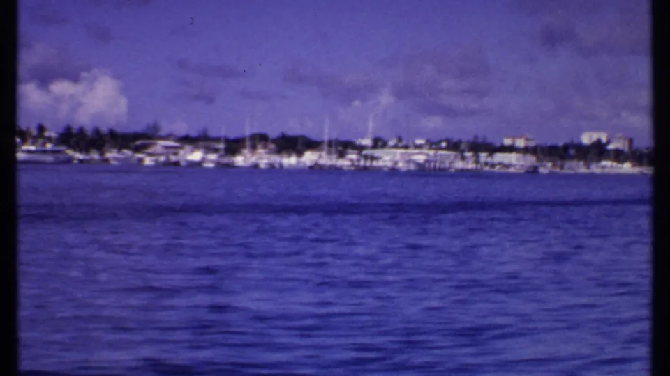 1969 Bahamas shoreline with docked boats and waterfront buildings across calm water, framed by visible 8mm film edges.