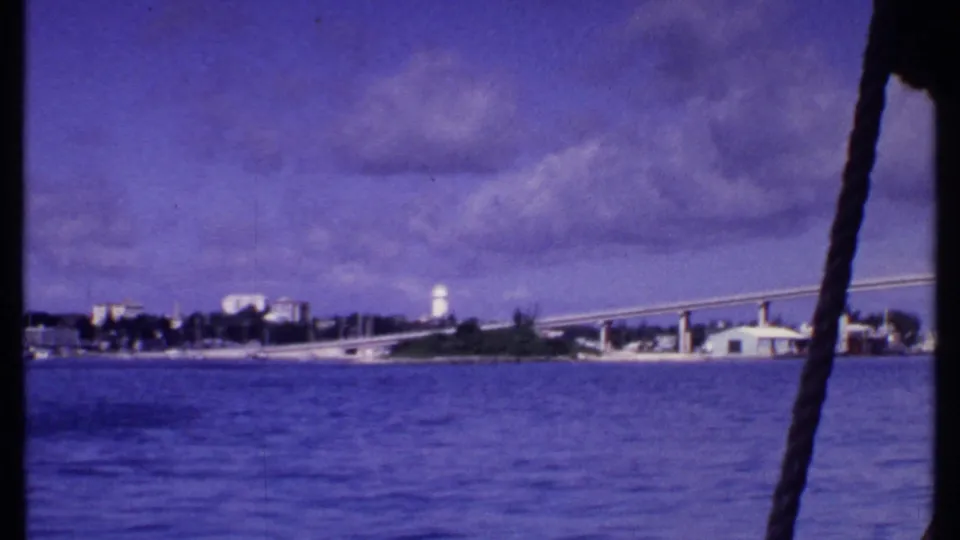 1969 Bahamas water view with a long bridge, distant shoreline buildings, clouds, and boat rigging visible at the right edge.