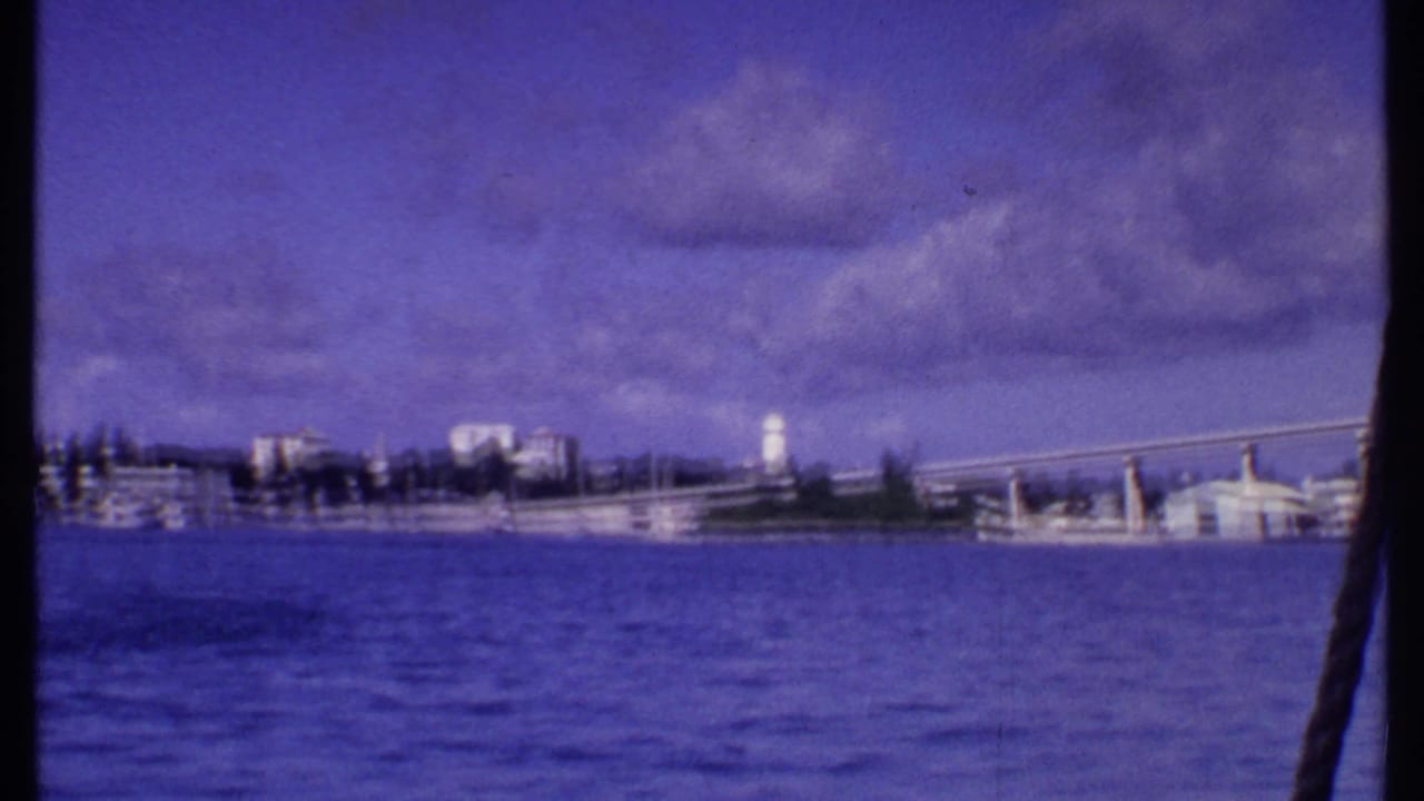 1969 Bahamas 8mm frame showing a waterfront from a boat, with blue water, low buildings, a bridge-like span, and a white tower.