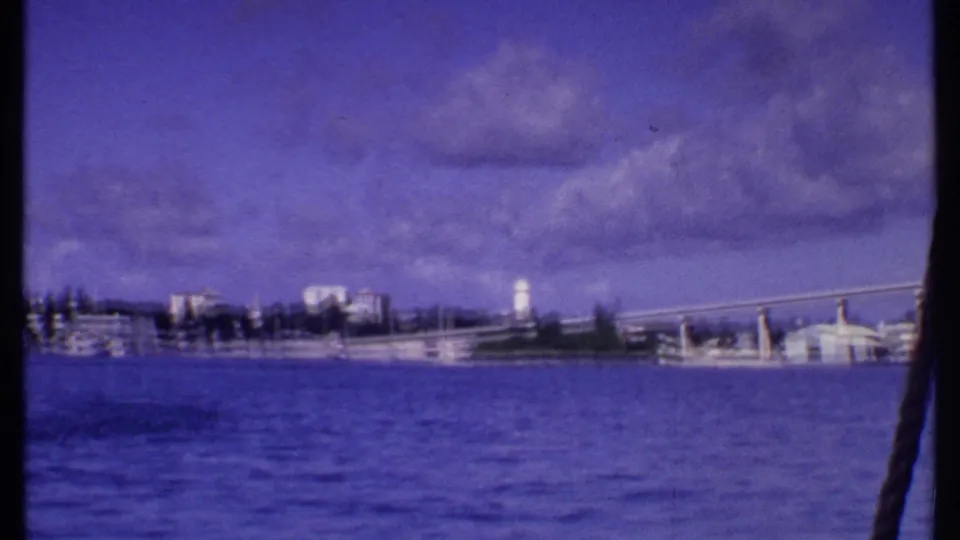 1969 Bahamas 8mm frame showing a waterfront from a boat, with blue water, low buildings, a bridge-like span, and a white tower.