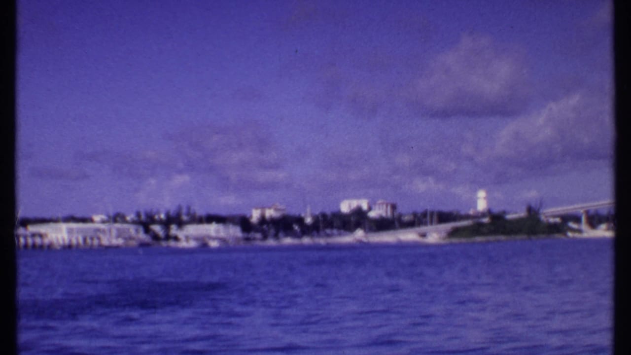 Distant shoreline with blue water in front, a white tower, low buildings, and a bridge or causeway at right.