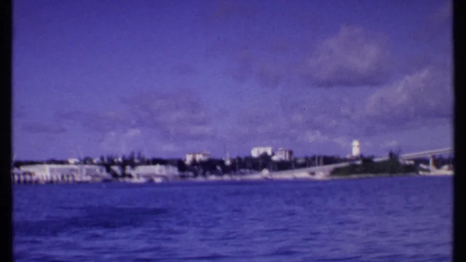 1969 Bahamas shoreline viewed from a boat, with low waterfront structures, a white tower, blue water, and a bridge or causeway at right.