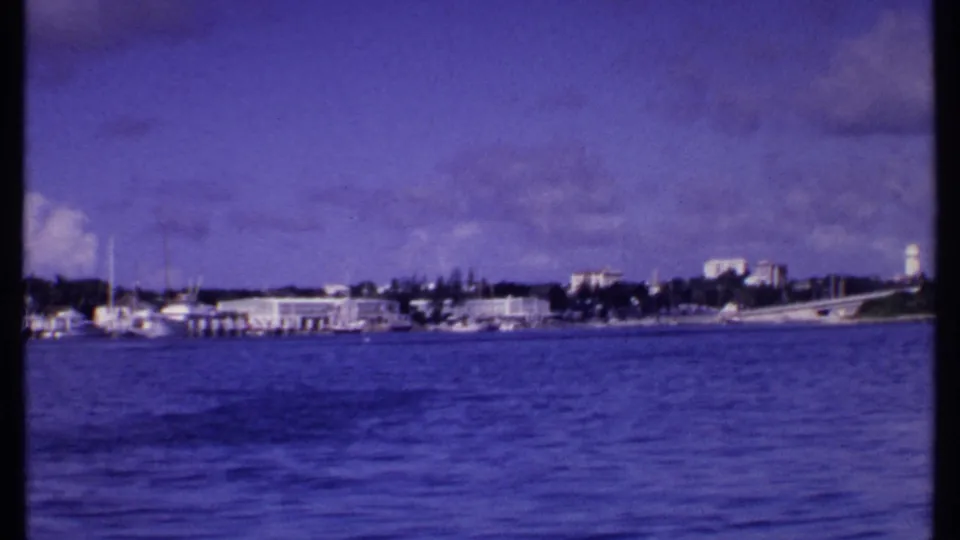 1969 Bahamas 8mm view from the water showing docked boats, low waterfront buildings, a white tower, and a bridge-like structure.