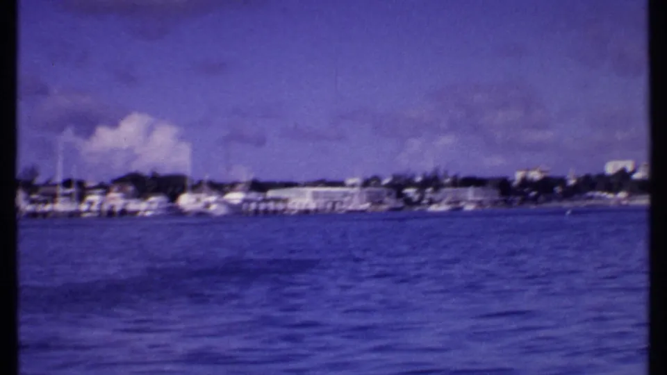1969 Bahamas boat-ride frame showing a waterfront with docked boats, low buildings, shoreline trees, blue water, and a cloudy sky.