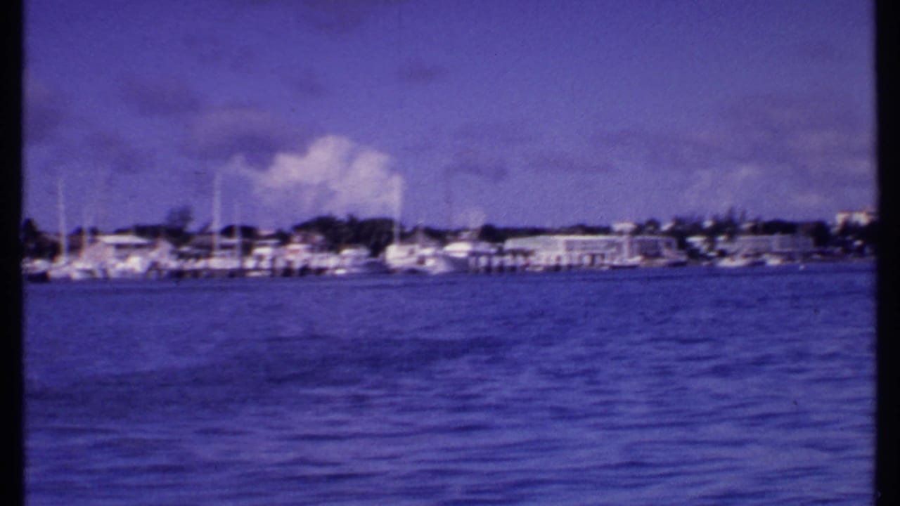 1969 Bahamas home-movie frame showing rippled harbor water, white sailboats at docks, and low buildings along the shore.