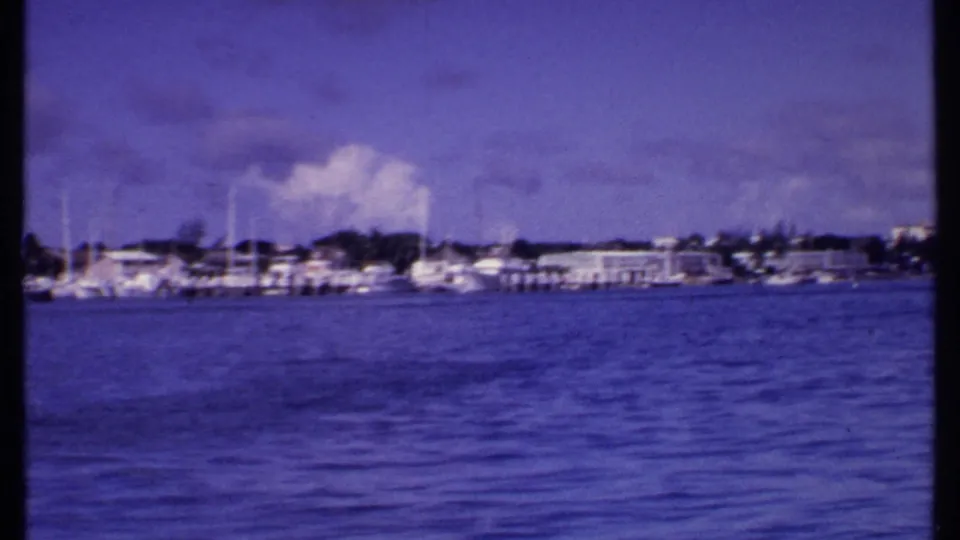 1969 Bahamas home-movie frame showing rippled harbor water, white sailboats at docks, and low buildings along the shore.