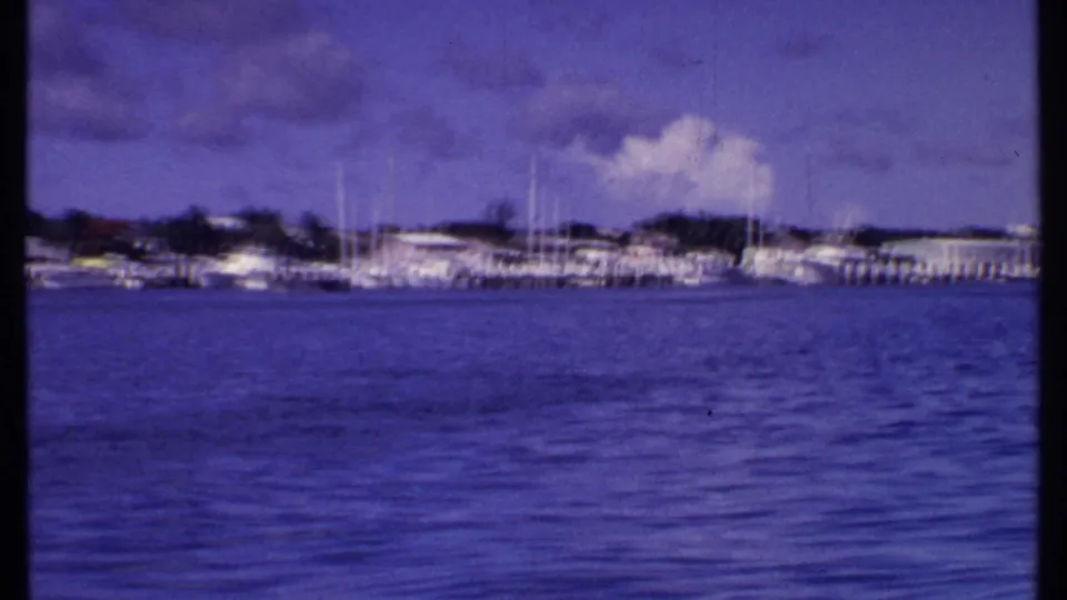 1969 Bahamas waterfront frame with a marina line, clustered white boats, tall masts, rippled water, and clouds above the shore.