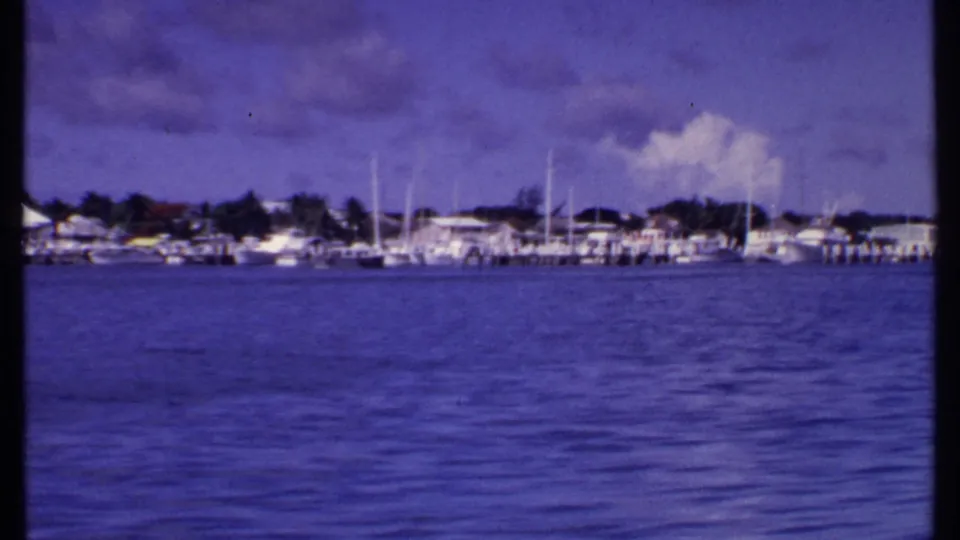 1969 Bahamas harbor frame showing distant sailboats, dock structures, low waterfront buildings, and a purple-blue aged-film cast.