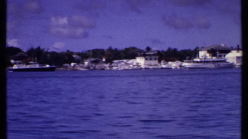 1969 Bahamas harbor frame with a dark-hulled boat on the water, nearby docks, white buildings, marina boats, and distant vegetation.