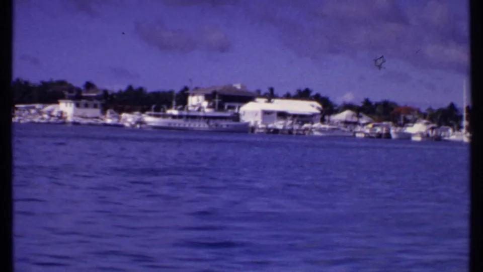 1969 Bahamas waterfront with a white yacht, dock structures, low buildings, and palms viewed from offshore.
