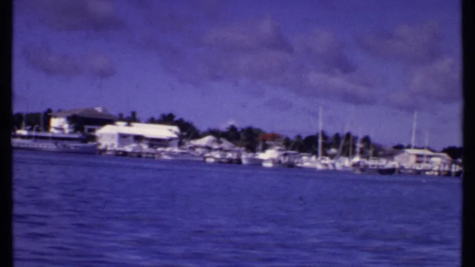 1969 Bahamas marina shoreline with docked boats, masts, low waterside buildings, palms, and purple-tinted sky.