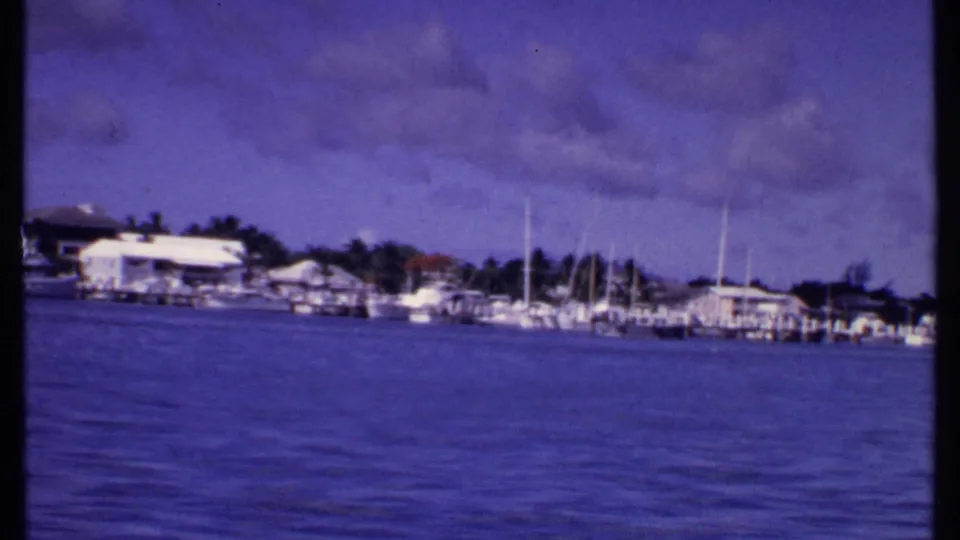 1969 Bahamas 8mm frame showing a distant marina across blue water with docked boats, masts, trees, and low buildings.