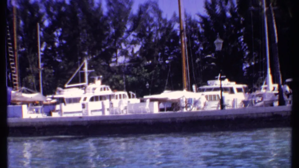 1969 Bahamas 8mm frame showing white yachts and sailboats along a marina dock, with blue-green water and tropical trees behind.