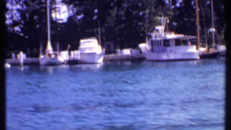 1969 Bahamas 8mm view of yachts and sailboats moored along a marina, seen from a moving boat across blue water.
