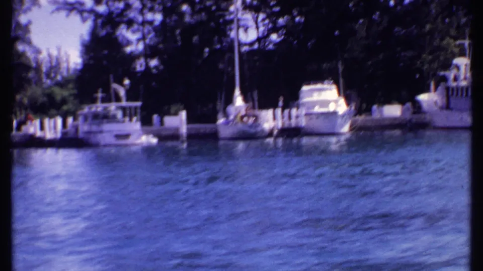 1969 Bahamas marina view with a small white boat passing near docked yachts, sailboats, white posts, palms, and shaded trees.