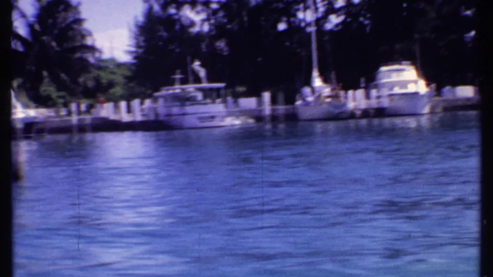 1969 Bahamas 8mm frame showing docked yachts, sailboats, palms, and bright blue marina water from a passing boat.