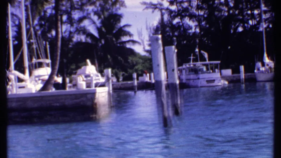 1969 Bahamas marina frame with sailboat masts, dock pilings, a white cabin boat, palms, and blue water filmed from a passing vessel.