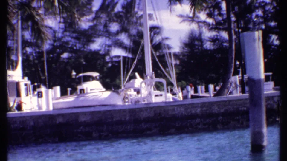 1969 Bahamas marina view with white sailboats, a motor yacht, concrete quay, palms, and distant people near the dock.