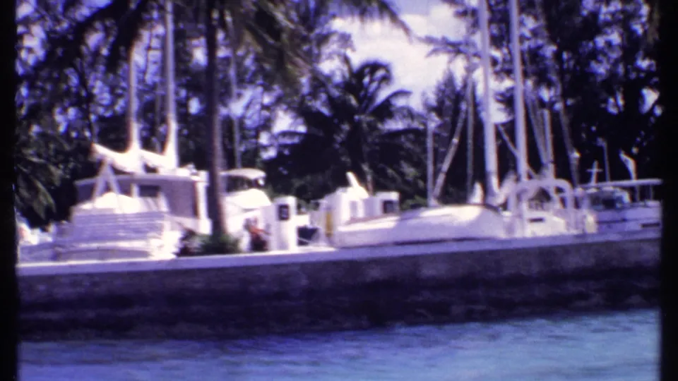 1969 Bahamas 8mm frame with white cabin boats, sail masts, palm trees, dock edge, and rippled marina water.