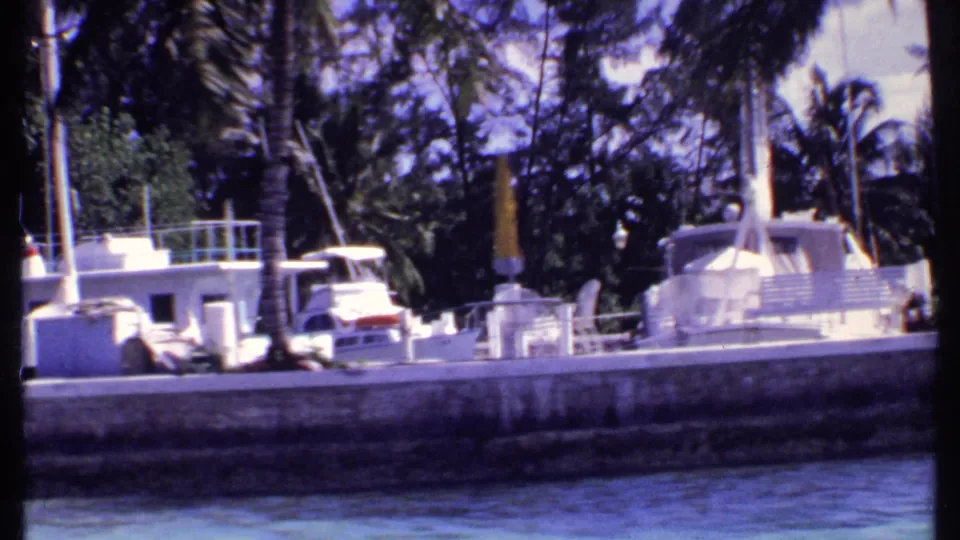 1969 Bahamas marina frame showing moored white boats, a small dockside building, palms, blue water, and distant people.