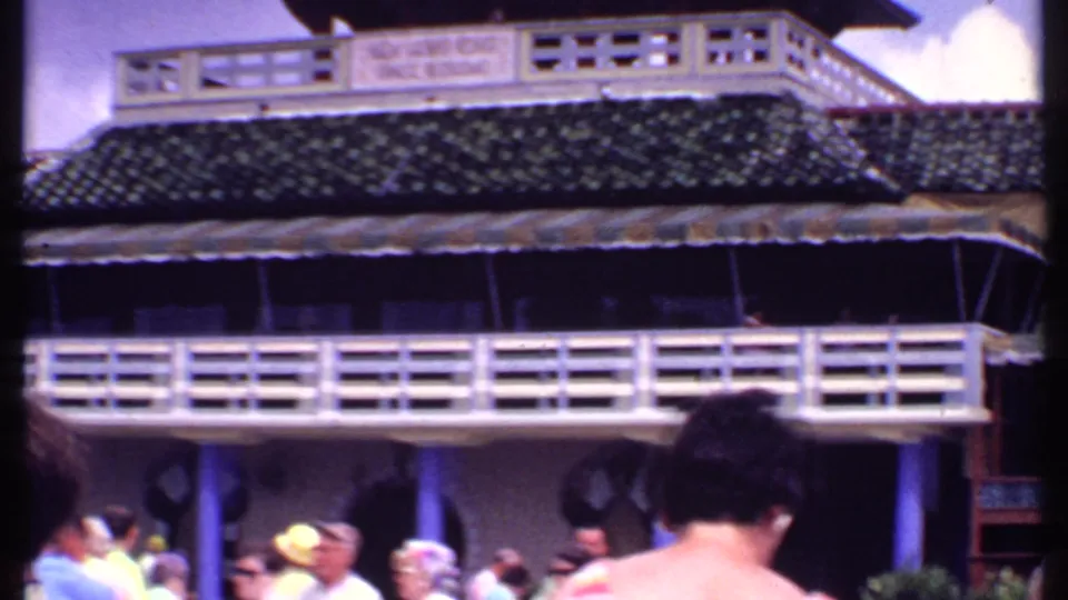 1969 Bahamas home-movie view of tourists walking beneath a pagoda-like building with white railings and a dark tiled roof.